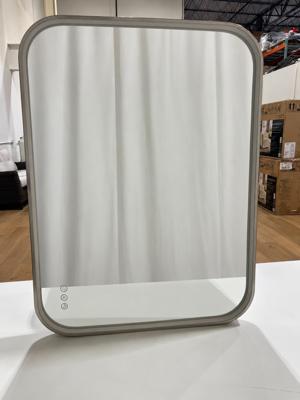 A rectangular wall mirror with rounded corners and a light gray frame, standing on a white surface in a warehouse-like setting with cardboard boxes visible in the background.