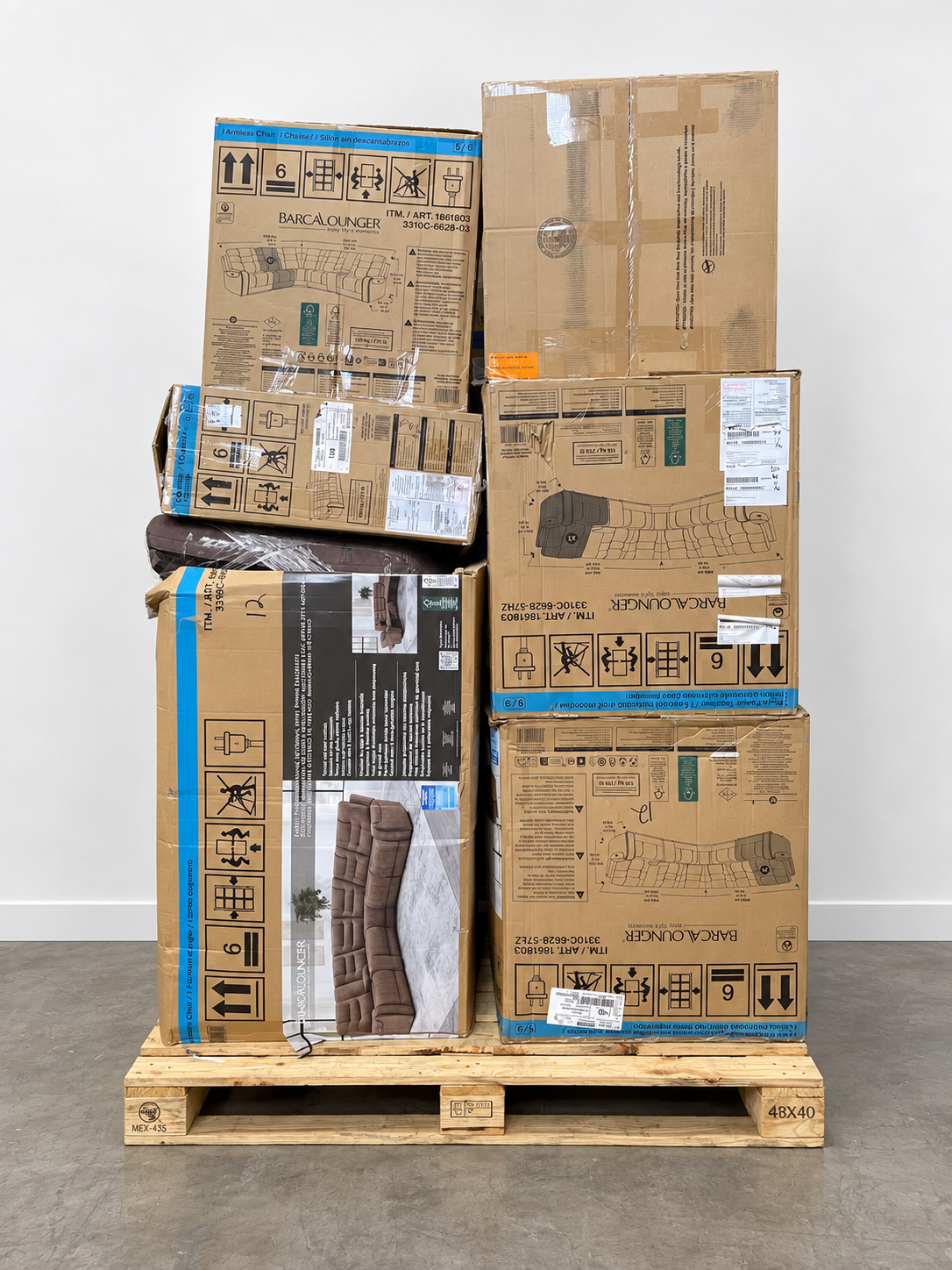 A tall stack of large cardboard shipping boxes on a wooden pallet against a plain light gray wall and concrete floor. Several boxes show printed packaging graphics and labels, including one box with a dark brown product photo.