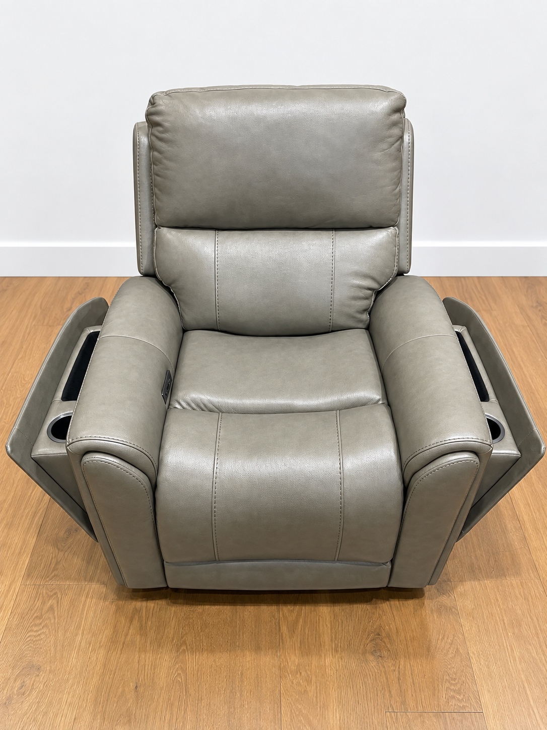 A gray faux leather recliner chair with side cup holders, shown from the front on a wooden floor against a white wall.