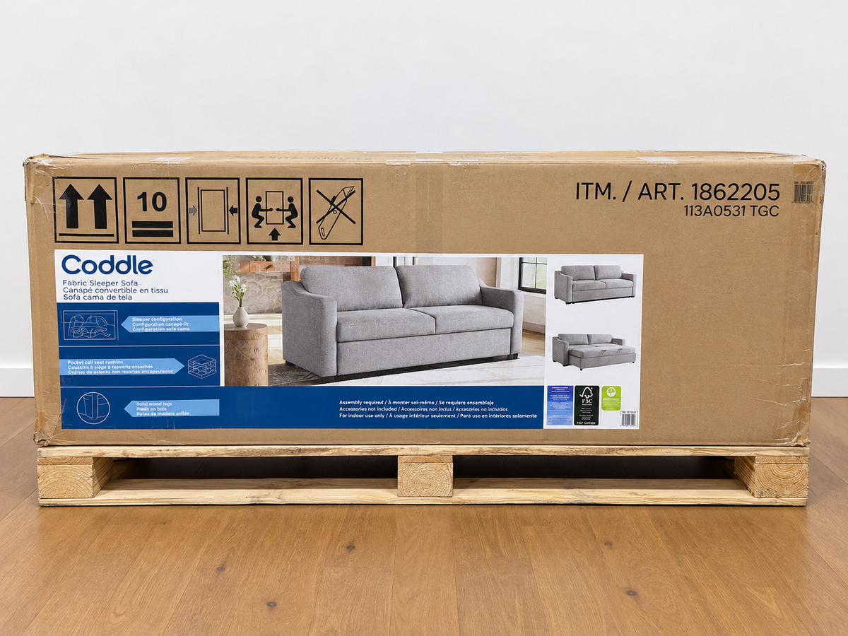 A large brown cardboard box for a Coddle sofa, displayed on a wooden pallet on a light wood floor against a white wall.