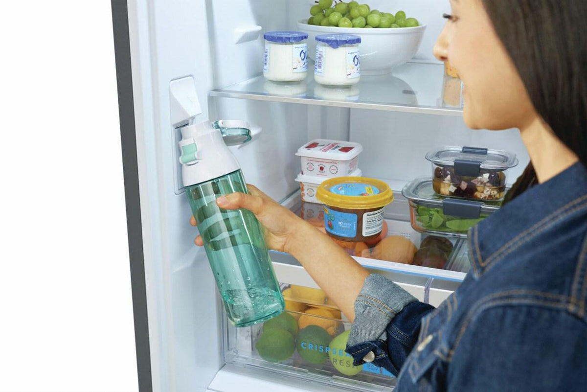 Close-up of a person filling a water bottle from the internal water dispenser, with the refrigerator stocked with food
