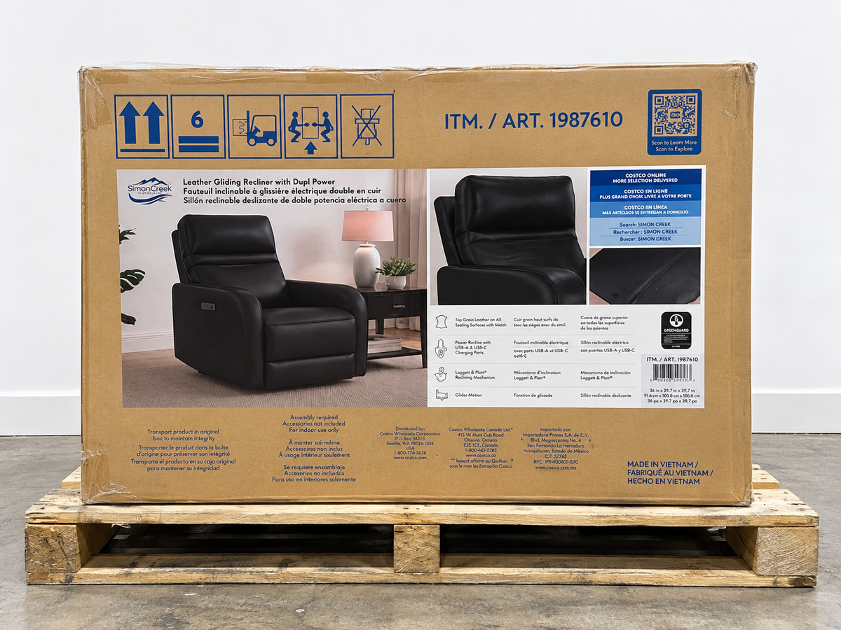 A large cardboard shipping box for a leather gliding recliner with dual power, photographed upright on a wooden pallet against a plain light gray wall and concrete floor. The box front shows product photos of the recliner, blue handling icons, item number ITM.