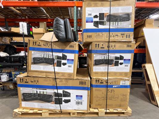A stack of large cardboard boxes containing black outdoor sectional sofas, secured with a black strap on a wooden pallet in a warehouse setting.