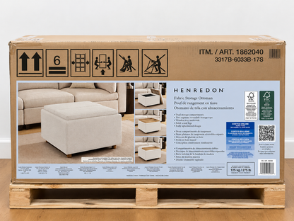 A large cardboard box for a Henredon fabric storage ottoman sits on a wooden pallet against a plain white wall. The box front shows product photos of the light beige ottoman, care instructions, barcode, and environmental icons.