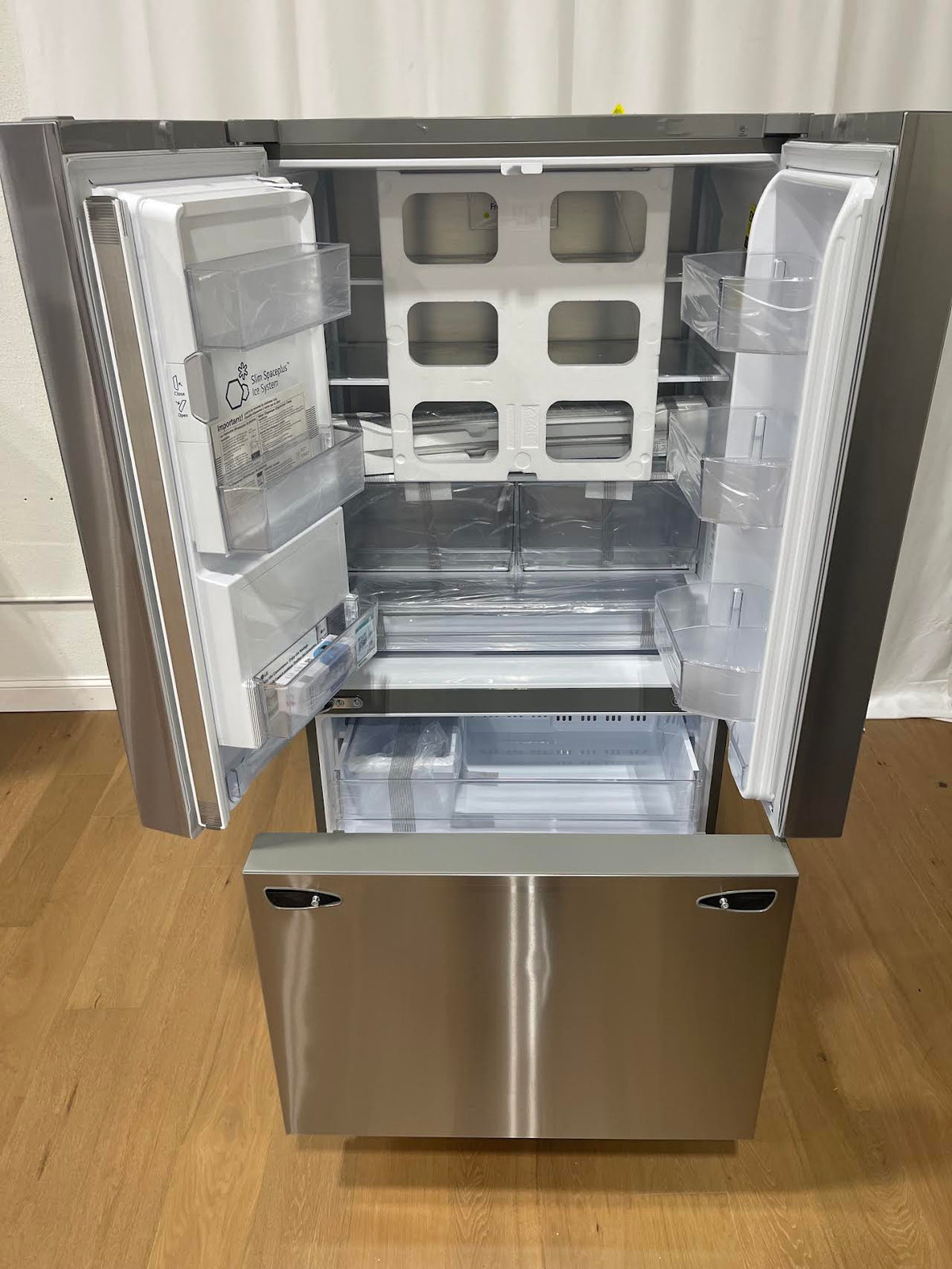 Refrigerator with both French doors open showing shelves and the Slim SpacePlus Ice System, still wrapped in factory plastic