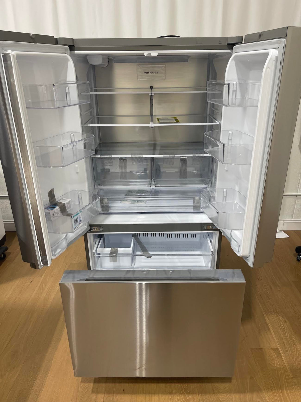 A stainless steel French door refrigerator with both upper doors open, showing the interior shelves and door bins, with the bottom freezer drawer pulled down. 