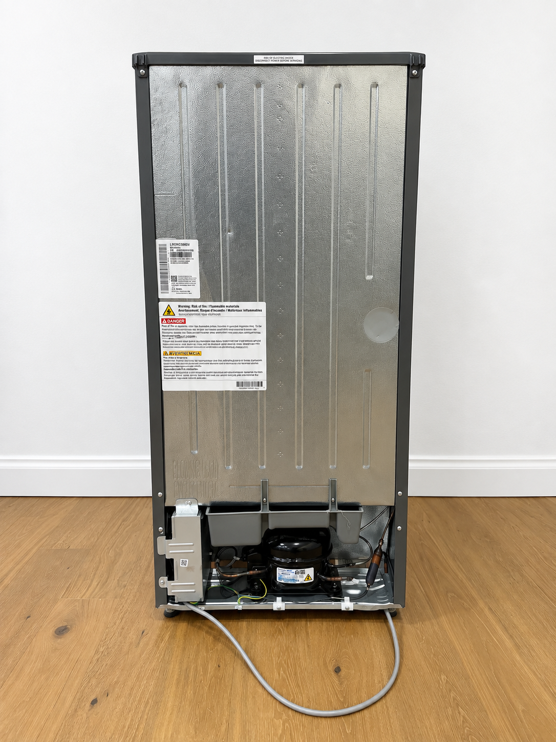 The back view of a tall stainless steel refrigerator or freezer, showing the vented metal back panel, warning labels, and exposed cooling components at the bottom.