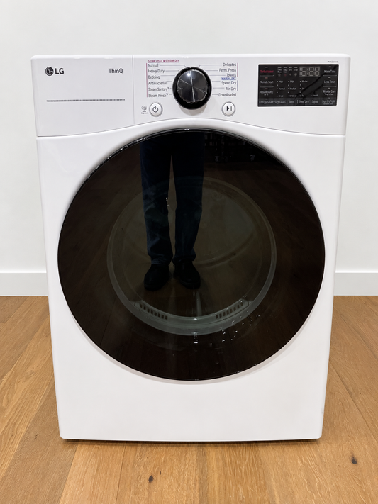 An LG ThauQ front-load washing machine in white, standing on a light wood floor against a white wall. The circular door has a dark reflective interior showing a reflection of a person's legs and shoes, which are incidental to the photo.