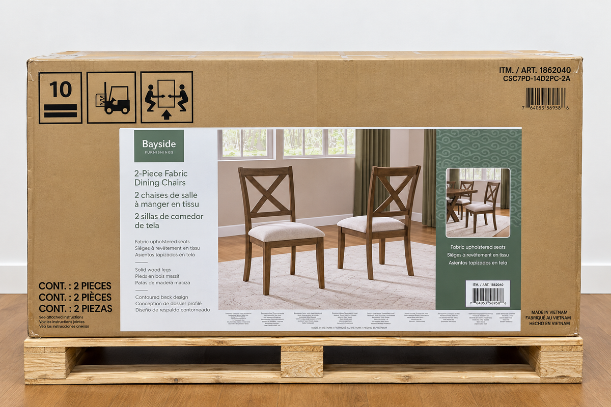 A large brown cardboard box for a Bayside 2-piece fabric dining chair set, displayed on a wooden pallet against a white wall and wood floor. The packaging features product images of the wooden chairs with off-white fabric seats.