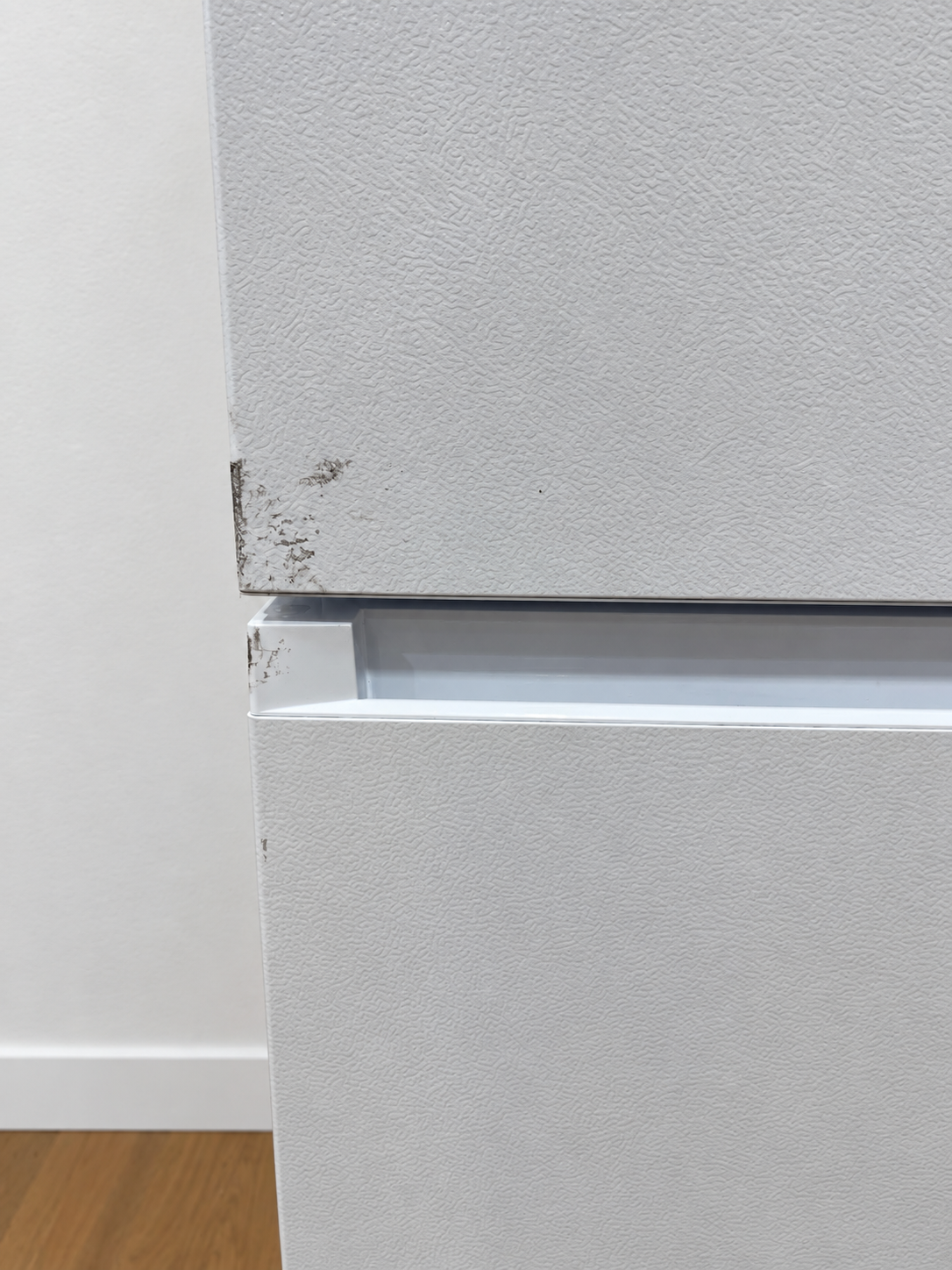 Close-up of the seam between the top and bottom drawers of a white textured refrigerator, showing some scuffs and dirt on the edge.