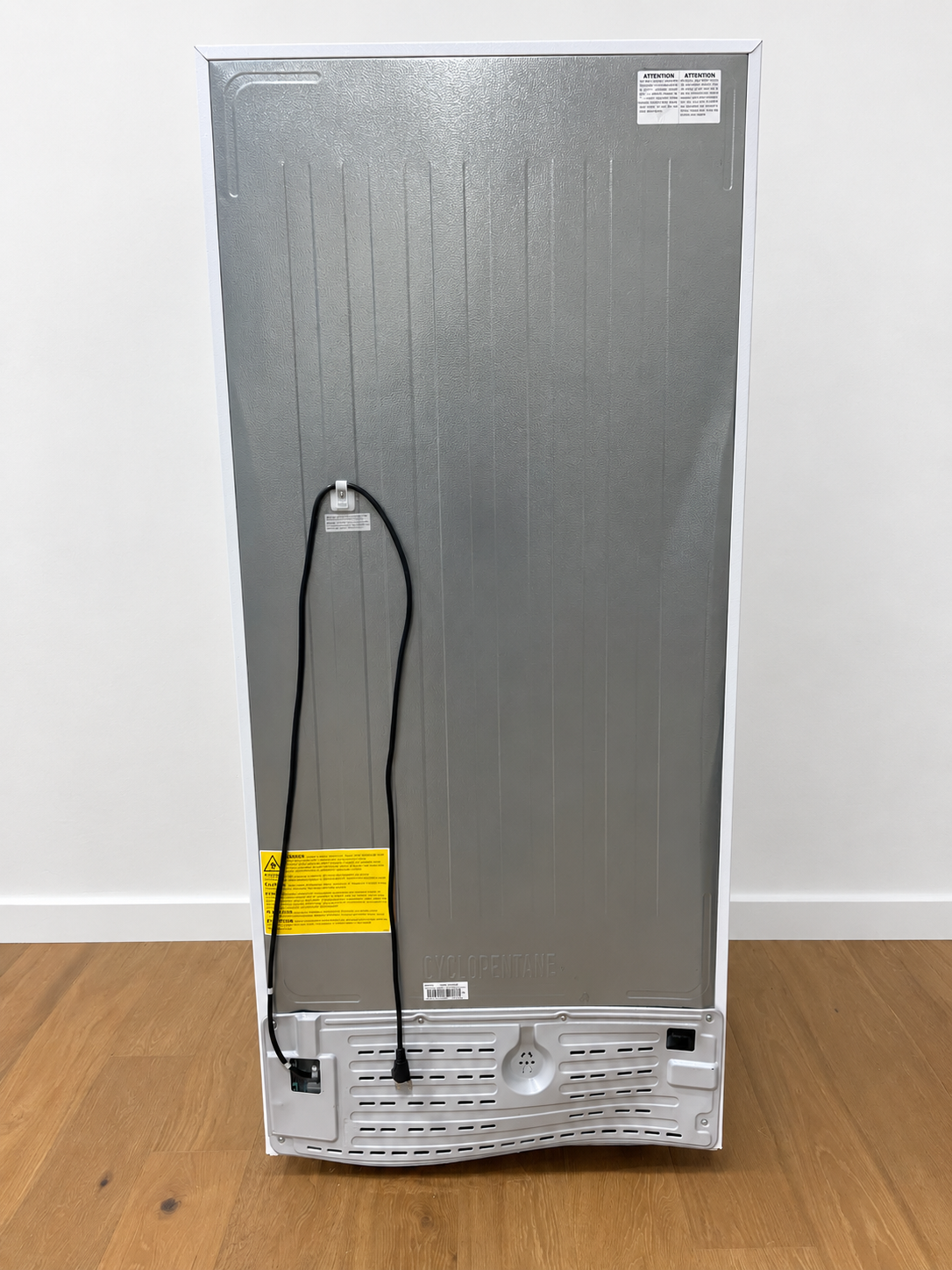 The back view of a tall, silver appliance, likely a refrigerator or freezer, standing on a light brown wooden floor against a white wall. The back panel is metallic silver with a black power cord hanging down, a yellow warning label, and showing a damaged white bottom grille.