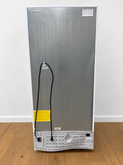 The back view of a tall, silver appliance, likely a refrigerator or freezer, standing on a light brown wooden floor against a white wall. The back panel is metallic silver with a black power cord hanging down, a yellow warning label, and showing a damaged white bottom grille.
