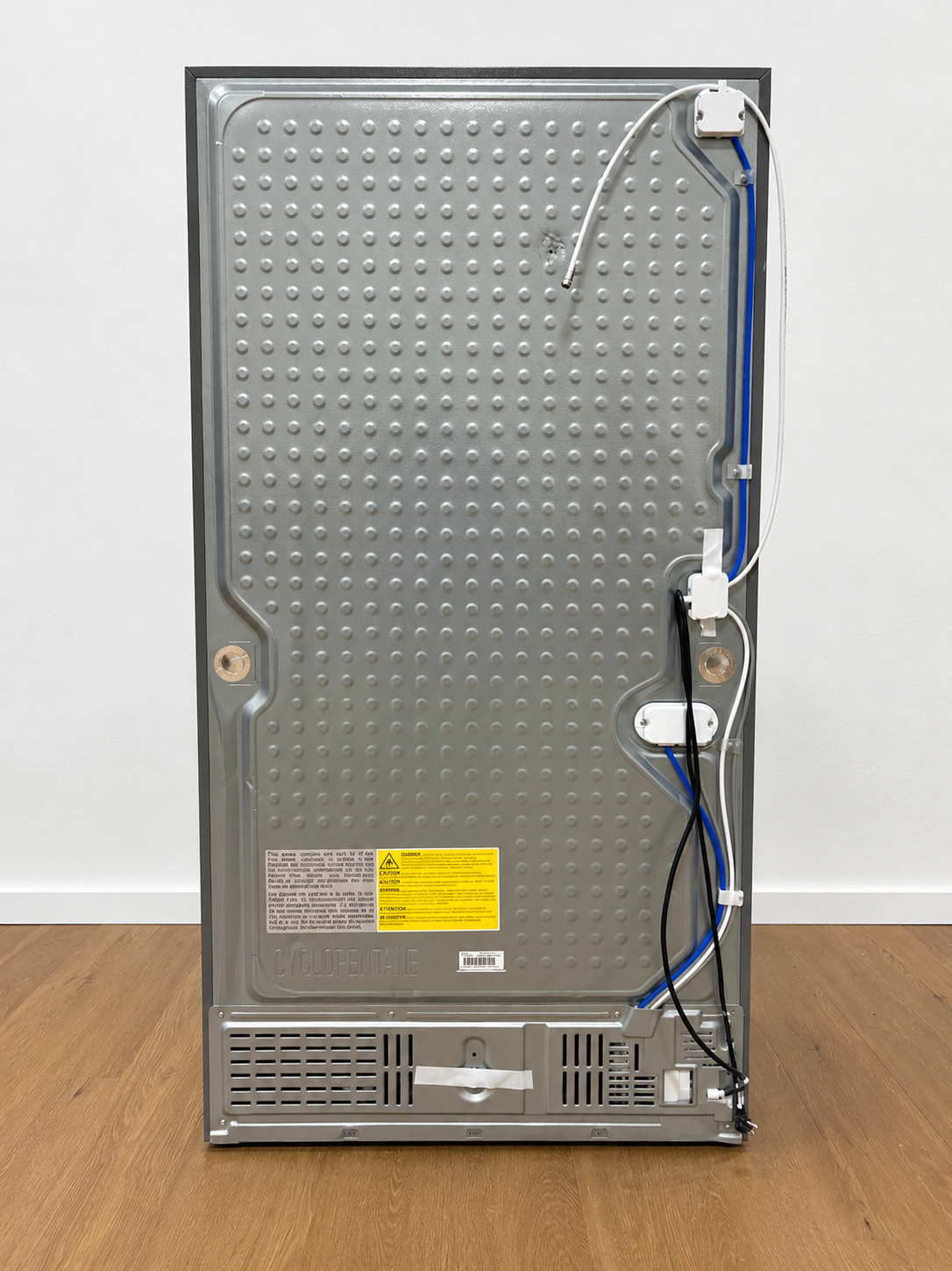 The back panel of a stainless steel appliance, likely a refrigerator or freezer. The back features a textured metallic surface with a yellow warning label, ventilation grilles, and various electrical wires and connectors.