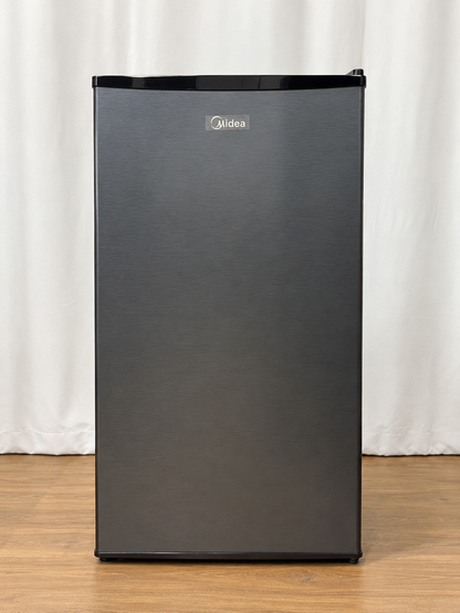 A Midea mini refrigerator with a brushed black stainless steel front and black top and bottom trim, standing on a wooden floor in front of a white curtain backdrop.