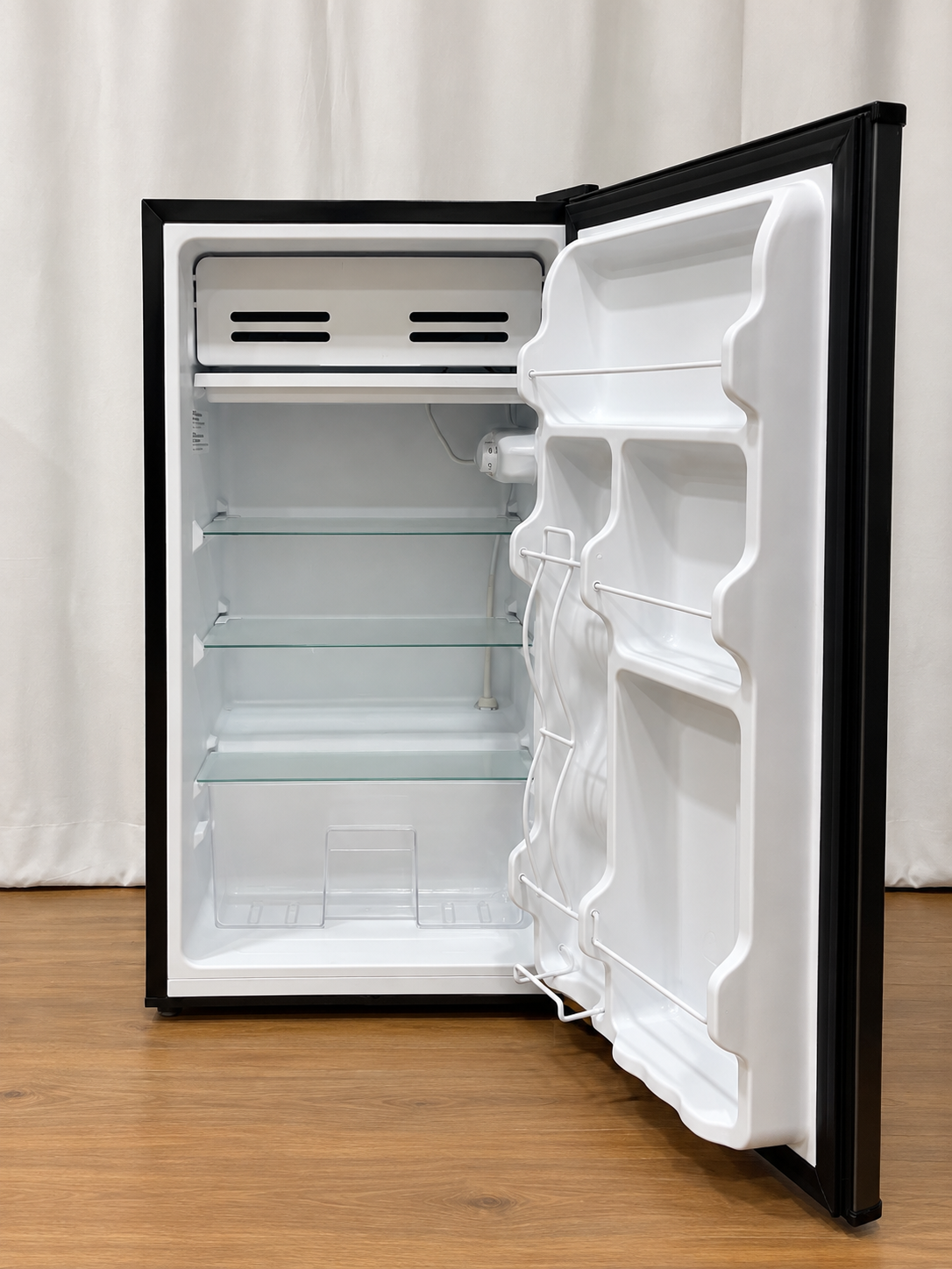 A compact mini refrigerator with a black exterior and white interior, shown with the door open on a wooden floor against a white curtain backdrop.