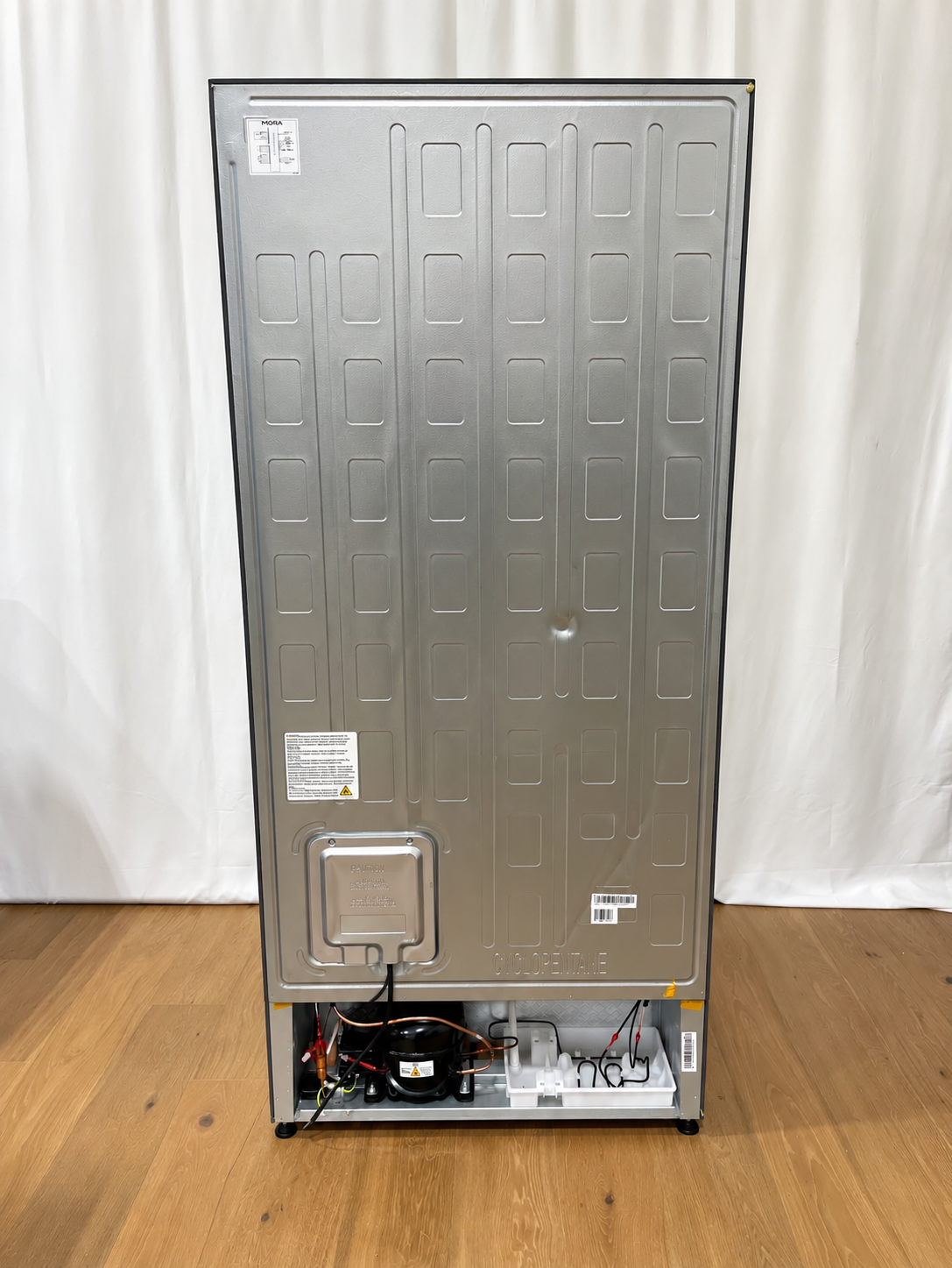 The back view of a tall stainless steel refrigerator or freezer, showing the rear panel, stickers, and the exposed cooling compartment with wires and compressor at the bottom.