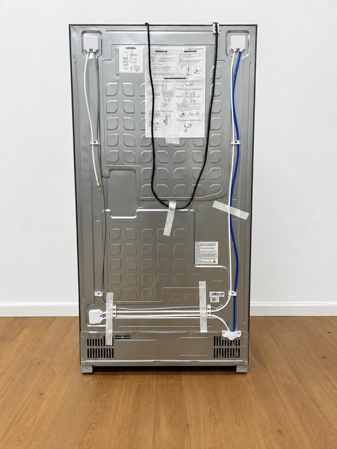 The back view of a stainless steel refrigerator, showing the internal wiring, cables, and printed technical labels on the back panel.