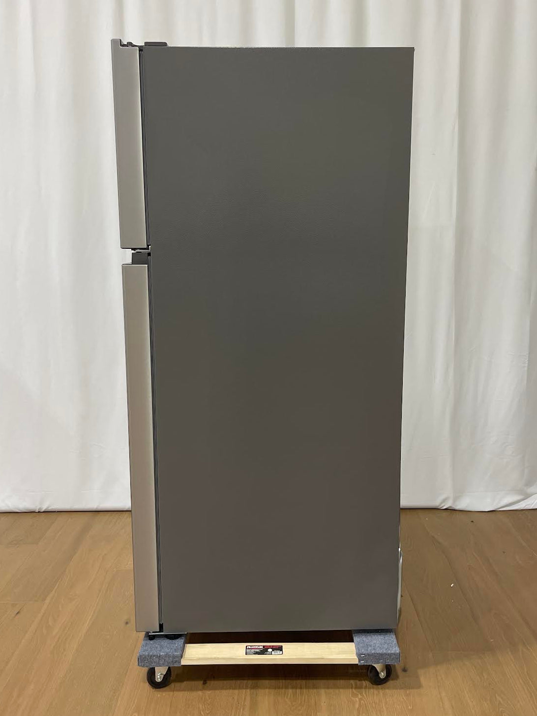 A tall, gray refrigerator with a top freezer compartment, shown standing on a wheeled dolly with a wooden board on a light wood floor in front of a white curtain backdrop.