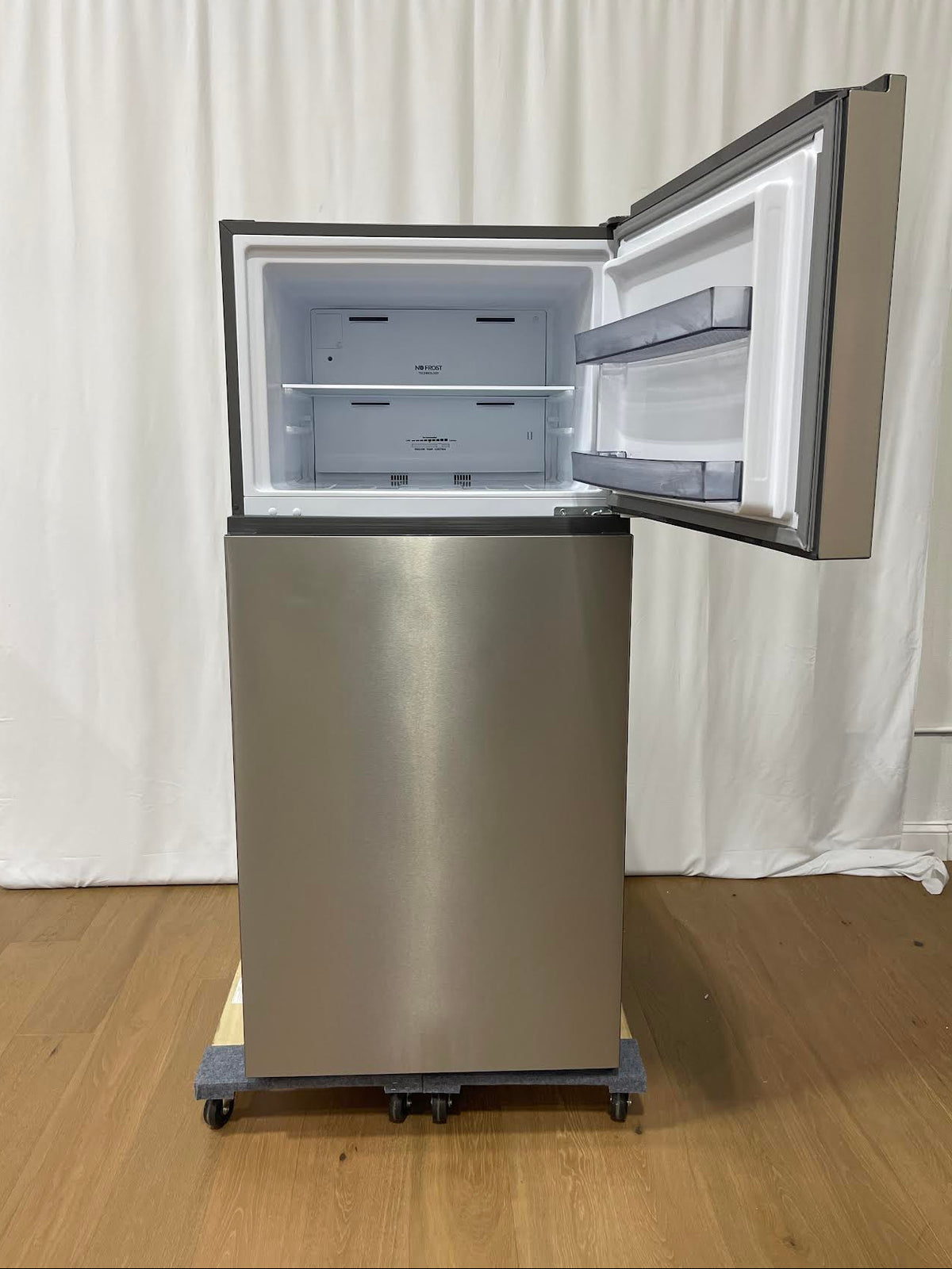 A stainless steel top-freezer refrigerator on wheels, shown with the freezer door open to reveal the empty interior, set against a white curtain backdrop on a wooden floor.