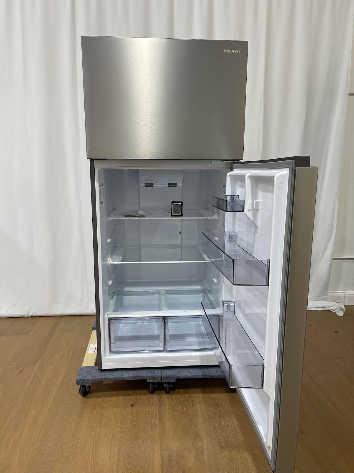 A stainless steel top-freezer refrigerator with its door open, showing a clean white interior with glass shelves and door bins, photographed on a wooden floor against a white curtain backdrop.
