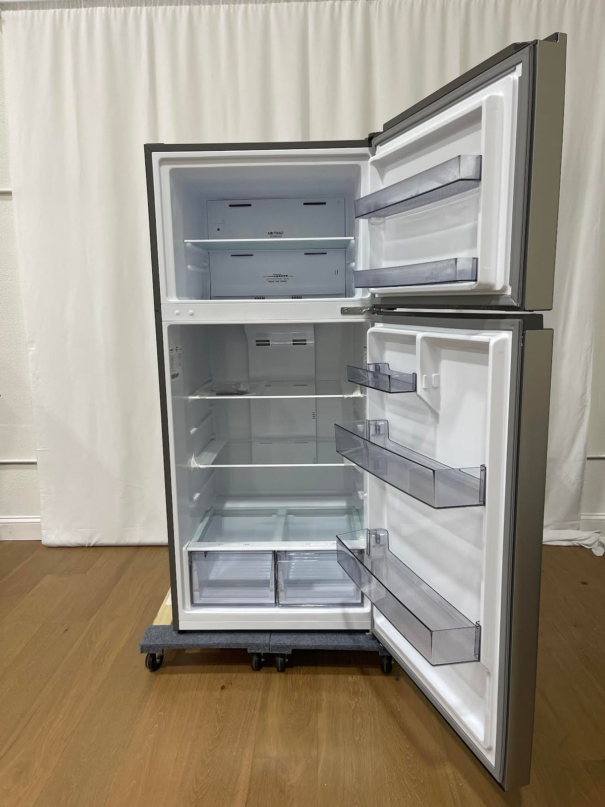 A top-freezer refrigerator with its doors open, showing a white interior with multiple shelves and translucent storage bins. The appliance has a dark gray exterior and sits on a wheeled dolly on a wooden floor in front of a white curtain backdrop.