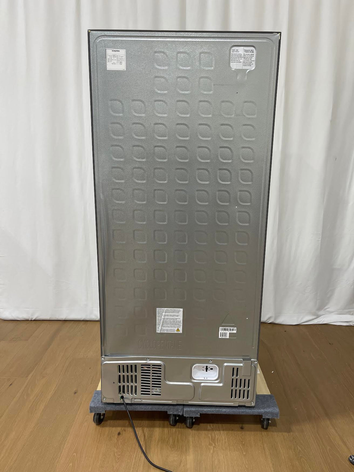 The back view of a stainless steel commercial refrigerator or freezer on a gray dolly with casters, set on a wooden floor against a white curtain backdrop.
