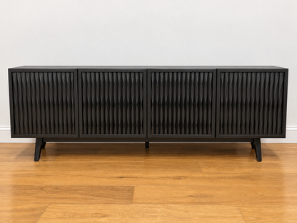 A modern black sideboard cabinet with four vertically slatted doors and tapered black legs, shown on a light wood floor against a plain white wall.