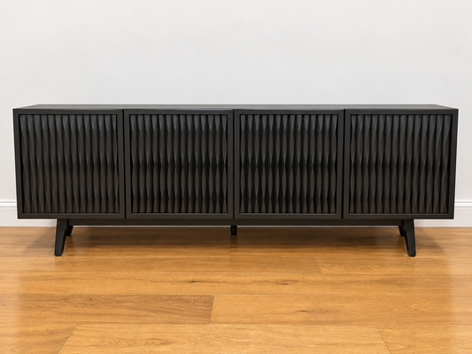 A modern black sideboard cabinet with four vertically slatted doors and tapered black legs, shown on a light wood floor against a plain white wall.