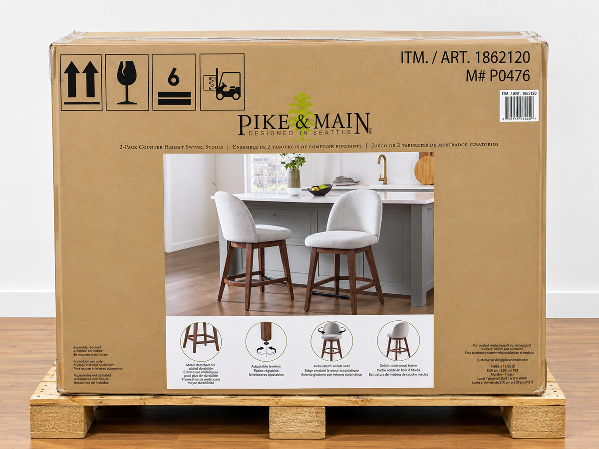 A large cardboard shipping box for Pike & Main bar stools sits on a wooden pallet in an indoor warehouse setting. The box features a photo of two white upholstered bar stools with dark wood legs, along with shipping symbols, item number ITM.
