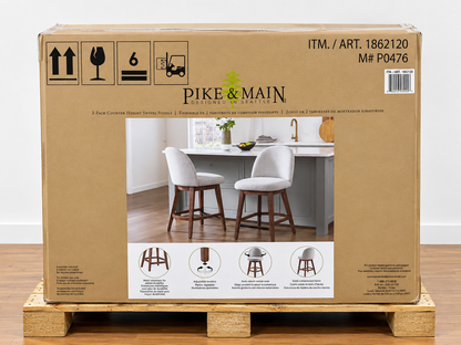 A large cardboard shipping box for Pike & Main bar stools sits on a wooden pallet in an indoor warehouse setting. The box features a photo of two white upholstered bar stools with dark wood legs, along with shipping symbols, item number ITM.