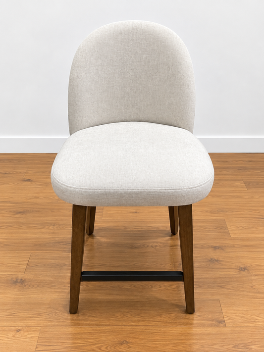 An upholstered dining chair with a light beige textured fabric seat and curved backrest, supported by dark brown wooden legs with a black metal footrest, set on a wooden floor against a white wall.