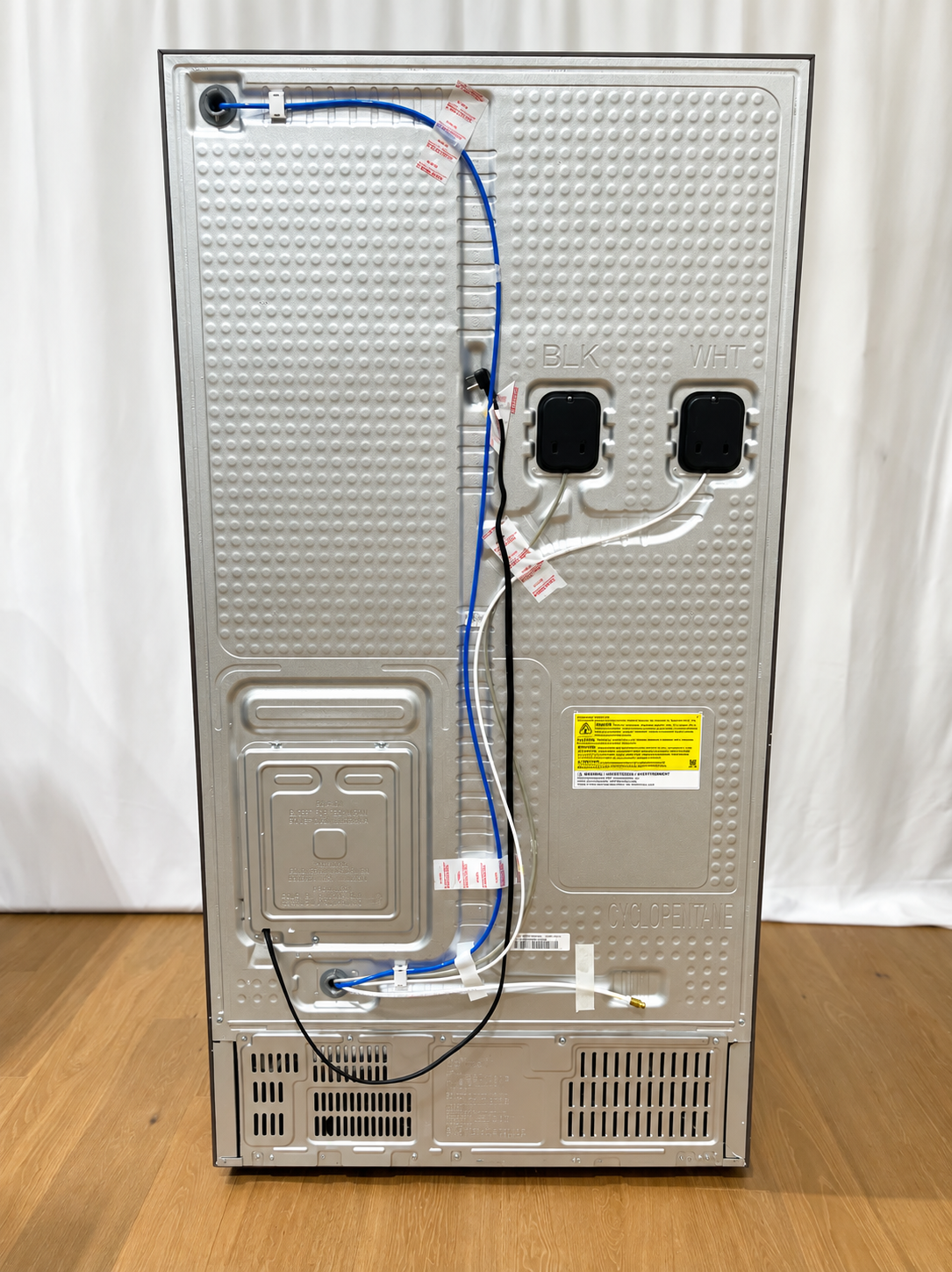 The back panel of a stainless steel refrigerator or freezer, showing the interior wiring, connection ports, vent grilles, and safety labels.
