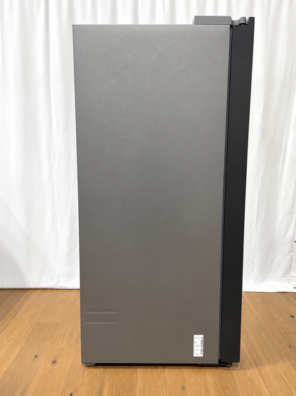 A tall, dark gray refrigerator with a black side panel, standing on a light wood floor in front of a white curtain backdrop.