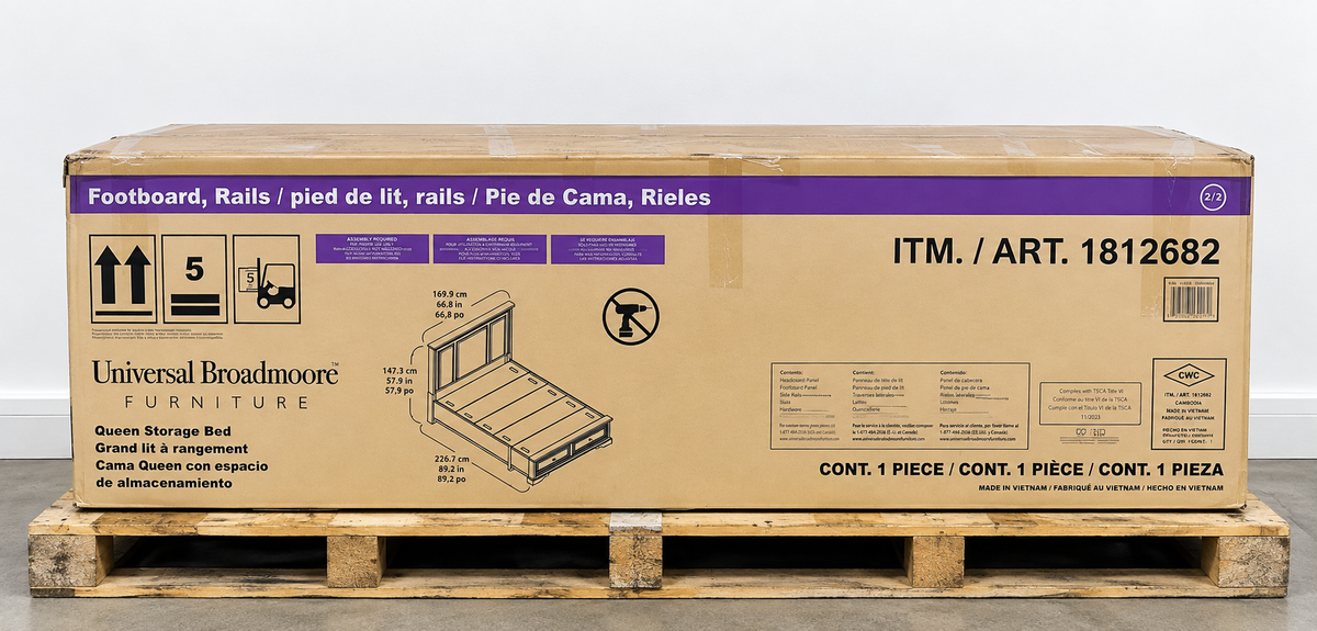A large Universal Broadmoore furniture box on a wooden pallet against a plain light gray wall. The box is tan cardboard with purple and black printed text and graphics, including the product name 'Footboard, Rails' and an illustration of a footboard frame.