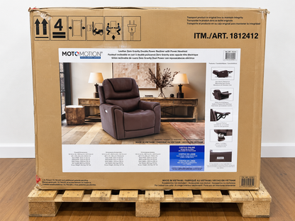A large cardboard box for a MOTOMOTION leather recliner, displayed on a wooden pallet against a white wall and wood floor. The box features a photo of a brown recliner and product specifications and diagrams.