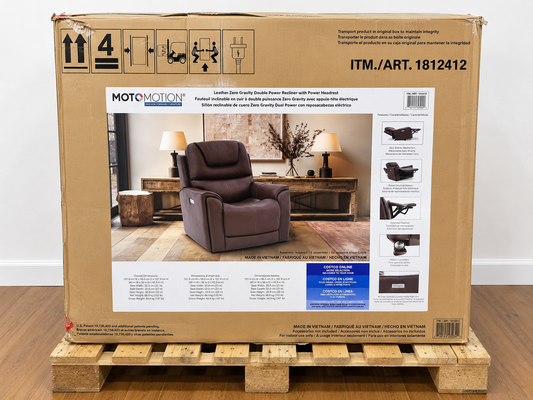 A large cardboard box for a MOTOMOTION leather recliner, displayed on a wooden pallet against a white wall and wood floor. The box features a photo of a brown recliner and product specifications and diagrams.